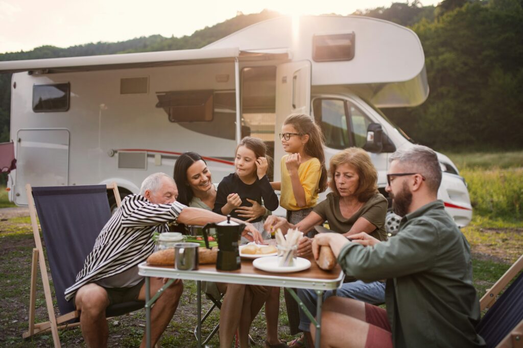 Multi-generation family sitting and eating outdoors by car, caravan holiday trip